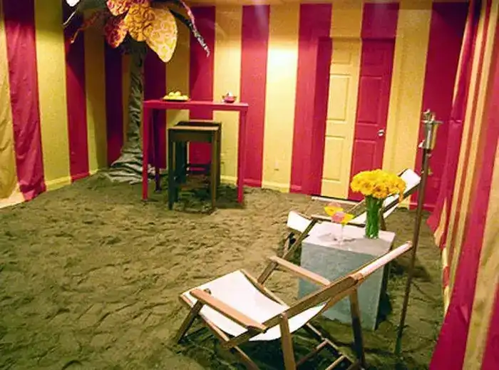 A room with grass-floor, red and yellow striped painted walls and a sunchair with a table standing by the right wall