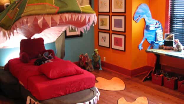 A bedroom mainly decorated in red. The bed is in the mouth of a fake dinosaur and the walls are ecorated with dinosaur-paintings.